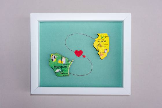Framed artwork with cut-out states of Wisconsin and Illinois connected by a heart on a teal background.