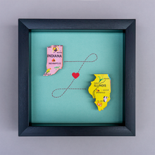  Framed artwork with pink Indiana and yellow Illinois shapes connected by a heart on a teal background.